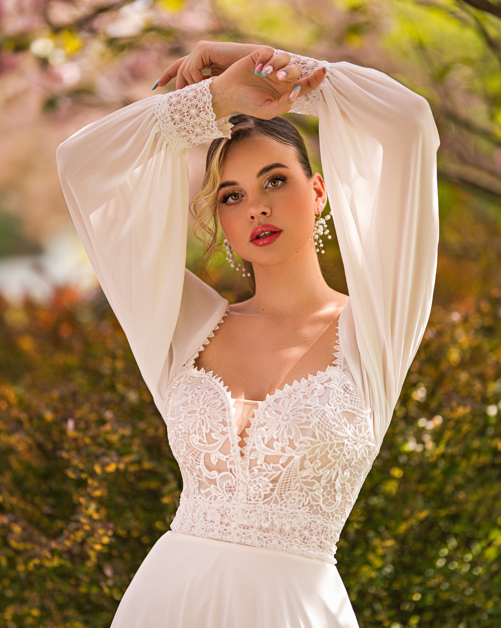 Portret kobiety w Olsztynie, z delikatnym, ślubnym rumieńcem na ustach i perłowymi kolczykami, pozującej w białej sukni z koronkowym dekoltem i bufiastymi rękawami na tle kwitnących drzew.