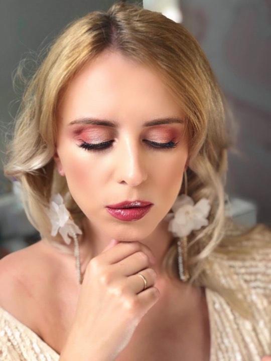 Portret kobiety z zamkniętymi oczami, prezentującej delikatny, różowy cień na powiekach, błyszczące usta i duże, białe kolczyki w kształcie kwiatów.