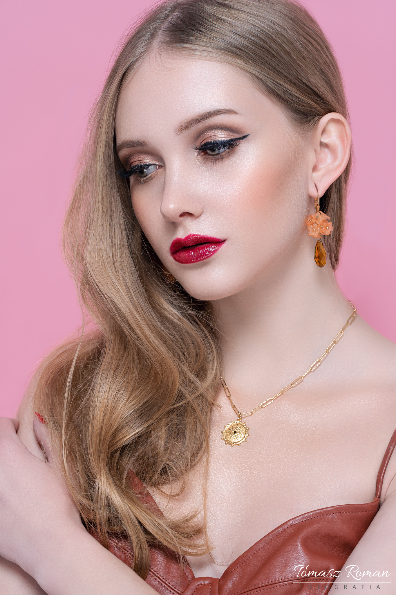 Portret kobiety z profesjonalnie wykonanym wieczorowym w odcieniach złota i burgundu, z precyzyjną kreską na powiece i błyszczącą szminką, uzupełniony o delikatną biżuterię na tle pudrowego różu.