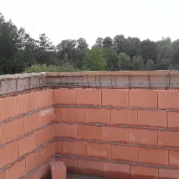 Wznoszone ściany z czerwonych bloczków ceramicznych, zbrojenie wieńca w drewnianym szalunku, widok na zalesione wzgórza w tle.