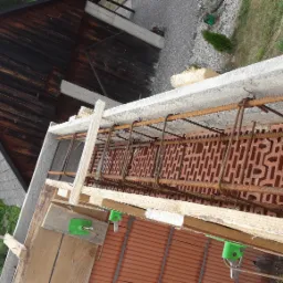 Wieniec żelbetowy w trakcie wykonywania, szalunek drewniany z zielonymi elementami mocującymi, widoczne zbrojenie oraz pustaki ceramiczne, w tle budynek gospodarczy z drewnianą elewacją.