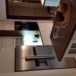 Nowoczesna kuchnia z jasnymi, drewnopodobnymi szafkami, stalową lodówką z dystrybutorem wody i blatem z dekoracjami. Na blacie widoczne szklanki i pleciona podkładka.