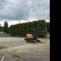 Żółto-szara koparka Volvo EC27C na terenie budowy z podsypką żwirową, w tle rząd wysokich tuj i pochmurne niebo.