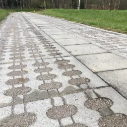 Długa, betonowa droga ułożona z dwóch rodzajów kostki: prostokątnej oraz ażurowej wypełnionej żwirem, prowadząca przez zielony teren w kierunku lasu w oddali.