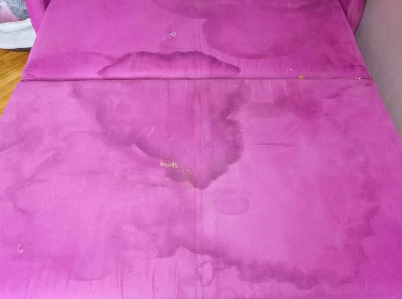 Fioletowa sofa z widocznymi, rozległymi plamami po zalaniu lub zabrudzeniach na siedzisku.