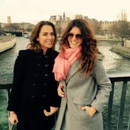 Dwie uśmiechnięte kobiety w płaszczach na tle Sekwany, Wrocław. Kobieta po prawej ma różowy szal i okulary przeciwsłoneczne, a w tle widać budynki.