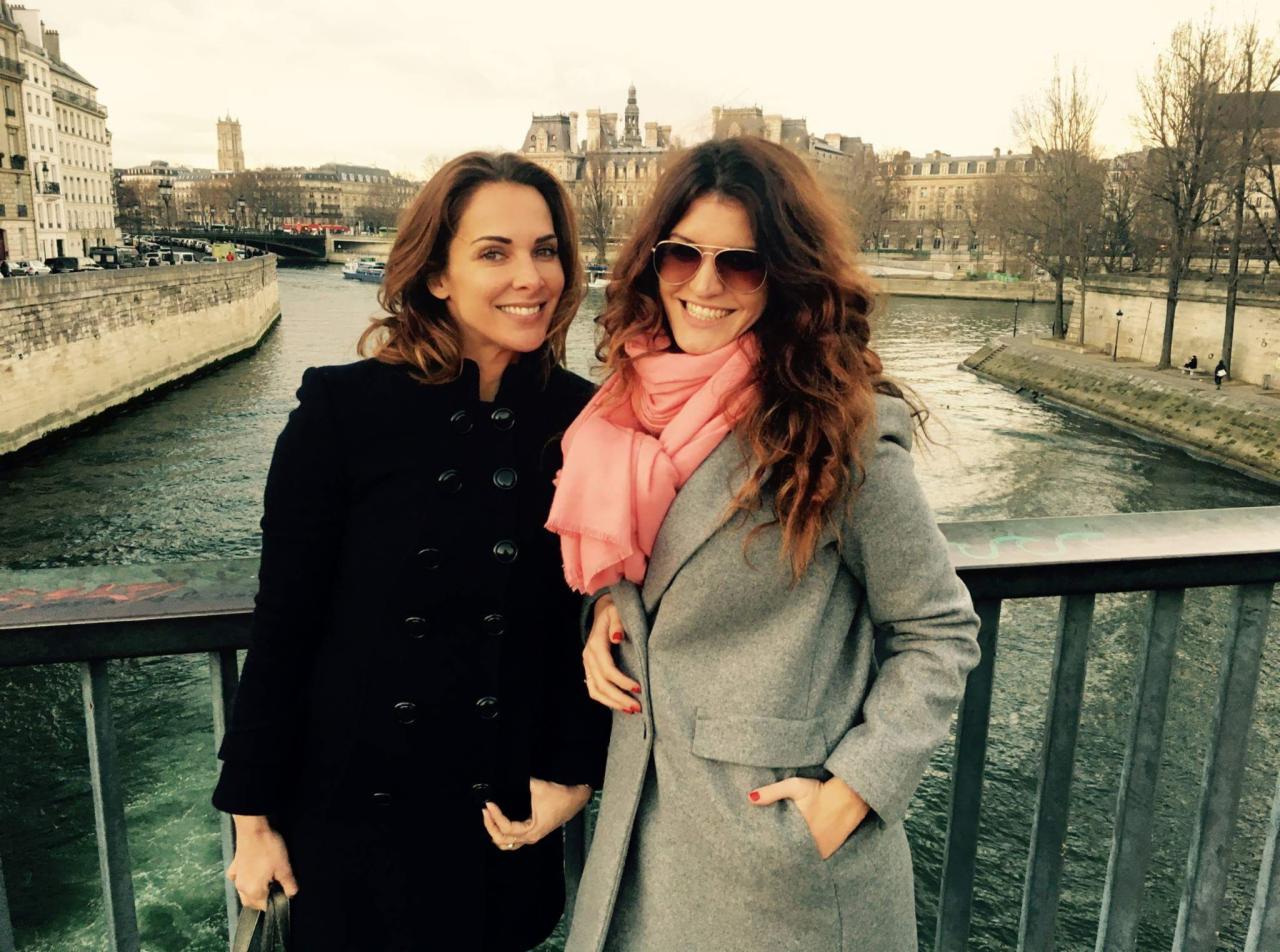 Dwie uśmiechnięte kobiety w płaszczach na tle Sekwany, Wrocław. Kobieta po prawej ma różowy szal i okulary przeciwsłoneczne, a w tle widać budynki.