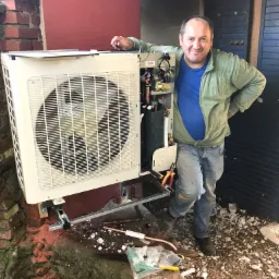 Mężczyzna w bluzie opiera się o zewnętrzną jednostkę klimatyzacji podczas montażu, widoczne narzędzia i fragment instalacji w trakcie podłączenia.