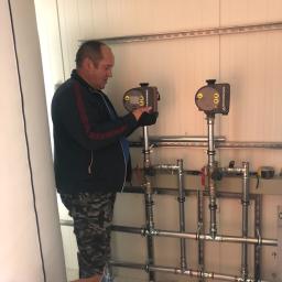 Instal-Term Mirosław Kaliś - Mężczyzna w czarnych rękawicach reguluje ustawienia na dwóch pompach obiegowych Grundfos X, zamontowanych na rurach instalacji grzewczej w pomieszczeniu o białych ścianach z metalowymi profilami.
