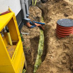 Instal-Term Mirosław Kaliś - Wykop z ułożoną pomarańczową rurą gazową zabezpieczoną żółtą taśmą ostrzegawczą GAZ, widoczny fragment instalacji przy budynku oraz pracownik w stroju roboczym.