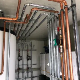 Instalacja grzewcza i hydrauliczna w kotłowni: miedziane i stalowe rury, biały zbiornik buforowy, pompy obiegowe Grundfos, manometry na szynie montażowej, widoczne zawory kulowe z czerwonymi rączkami.