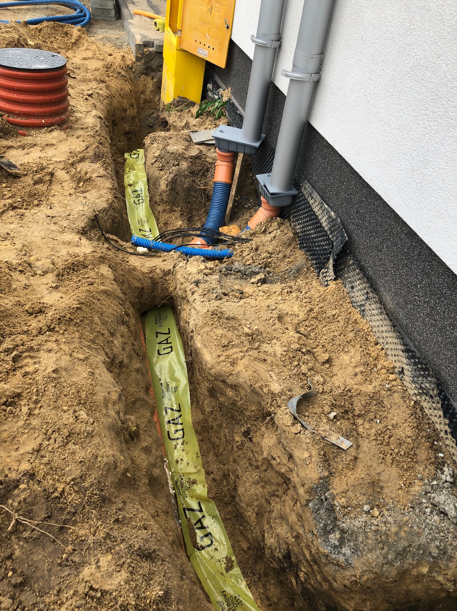 Wykop z ułożoną pomarańczową rurą gazową, zabezpieczoną żółtą folią ostrzegawczą z napisem GAZ, widok z góry na tle fundamentu budynku z orynnowaniem.