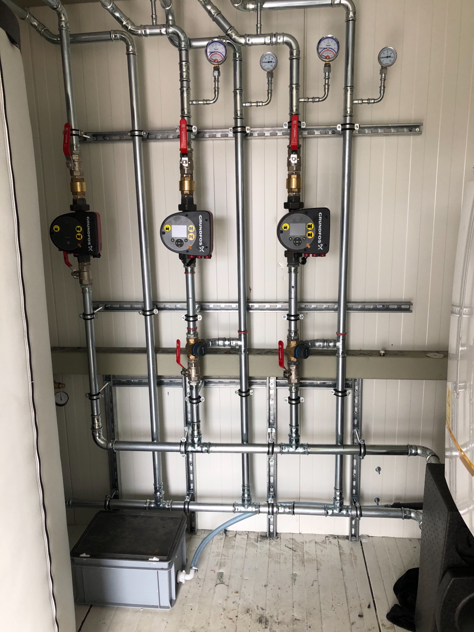 Instalacja z rur stalowych z manometrami i pompami obiegowymi Grundfos zamontowana na ścianie z paneli ściennych, widoczny zbiornik wyrównawczy.