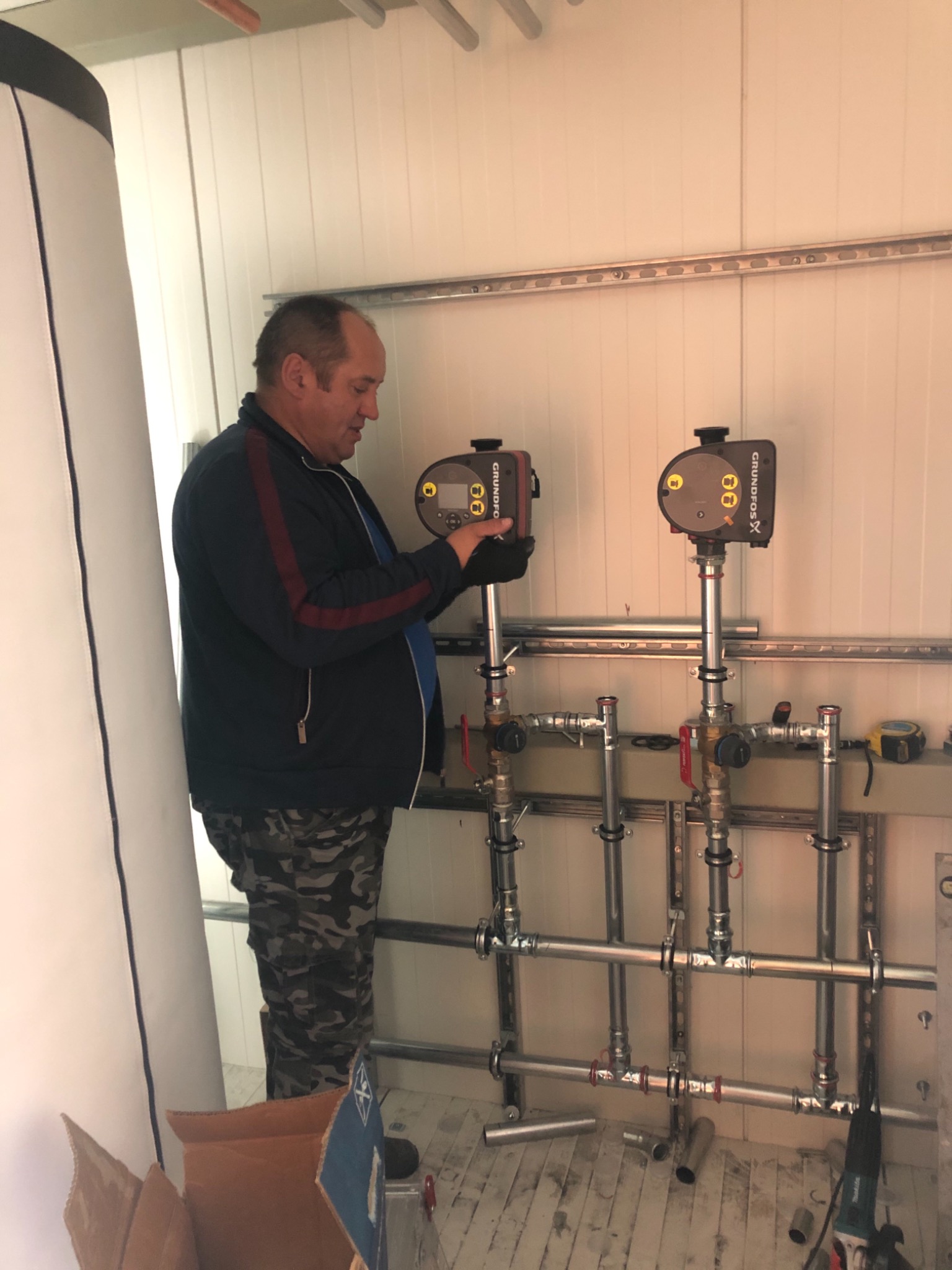 Mężczyzna w czarnych rękawicach reguluje ustawienia na dwóch pompach obiegowych Grundfos X, zamontowanych na rurach instalacji grzewczej w pomieszczeniu o białych ścianach z metalowymi profilami.