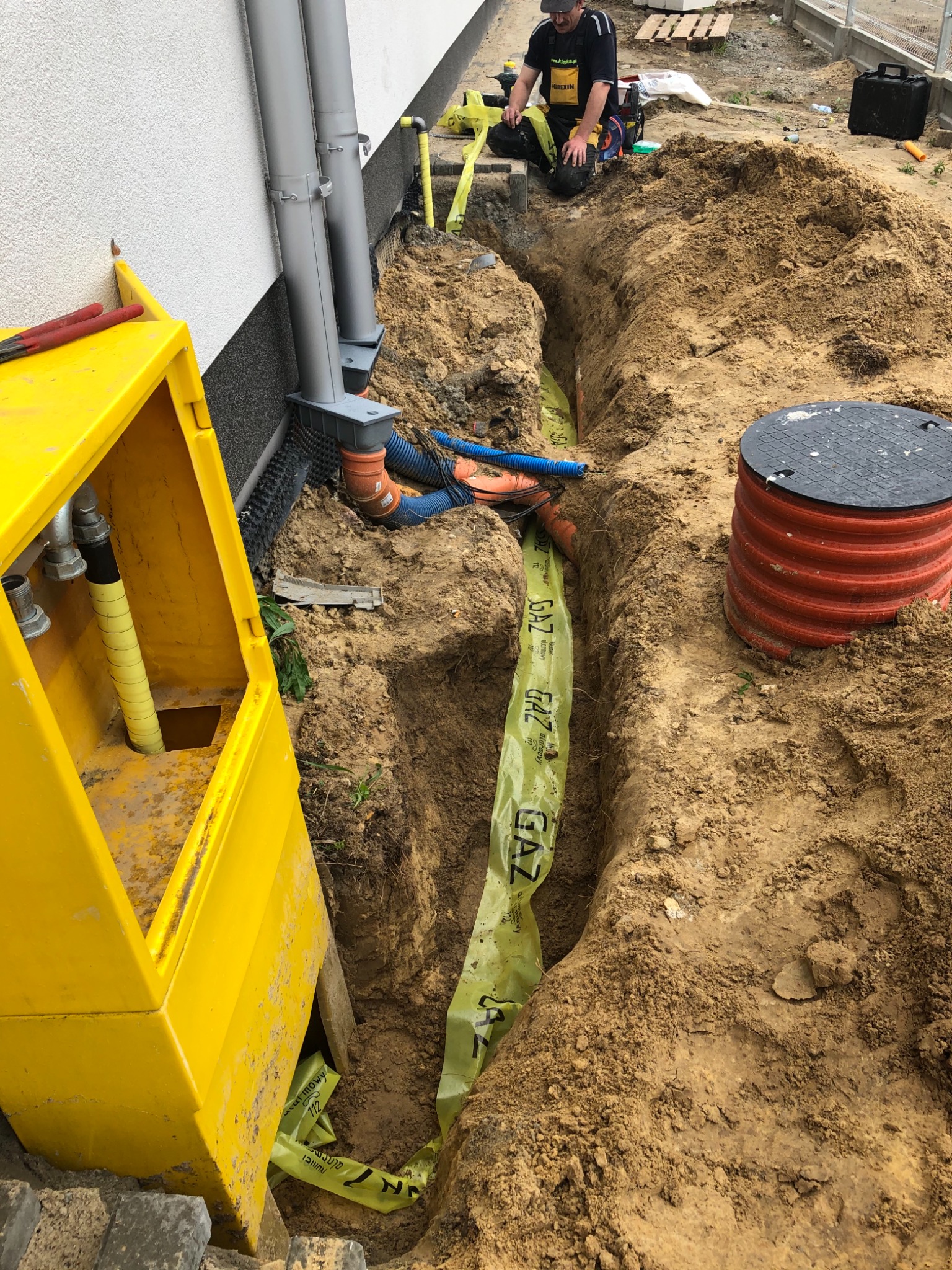 Wykop z ułożoną pomarańczową rurą gazową zabezpieczoną żółtą taśmą ostrzegawczą GAZ, widoczny fragment instalacji przy budynku oraz pracownik w stroju roboczym.