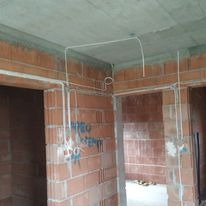 Surowy stan: Przewody elektryczne prowadzone po ścianach z czerwonej cegły i pod sufitem z betonu, widoczne puszki instalacyjne, wnętrze w trakcie budowy.