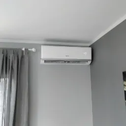 Biały klimatyzator Gree zamontowany na szarej ścianie w pokoju, obok okna z zasłoną i obrazu.