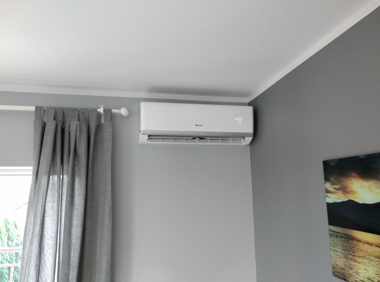 Biały klimatyzator Gree zamontowany na szarej ścianie w pokoju, obok okna z zasłoną i obrazu.