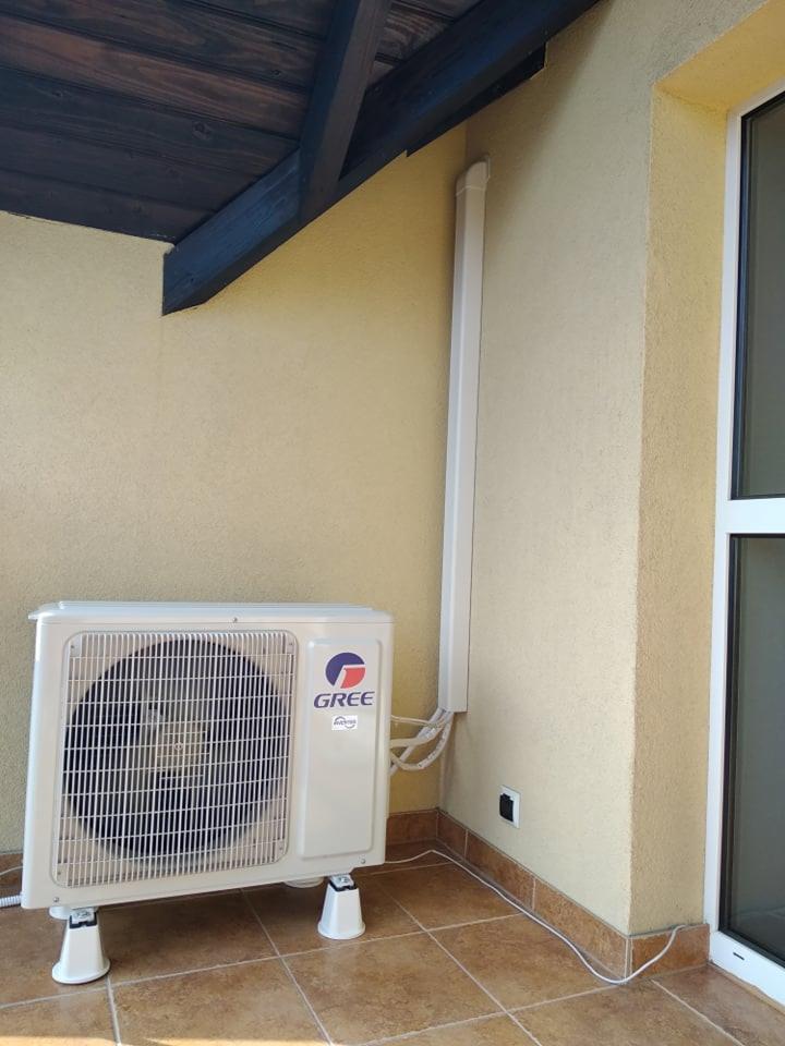 Zewnętrzna jednostka klimatyzatora marki Gree zamontowana na balkonie z kremową ścianą i brązowymi płytkami podłogowymi. Biała rura odprowadzająca skropliny biegnie wzdłuż ściany.
