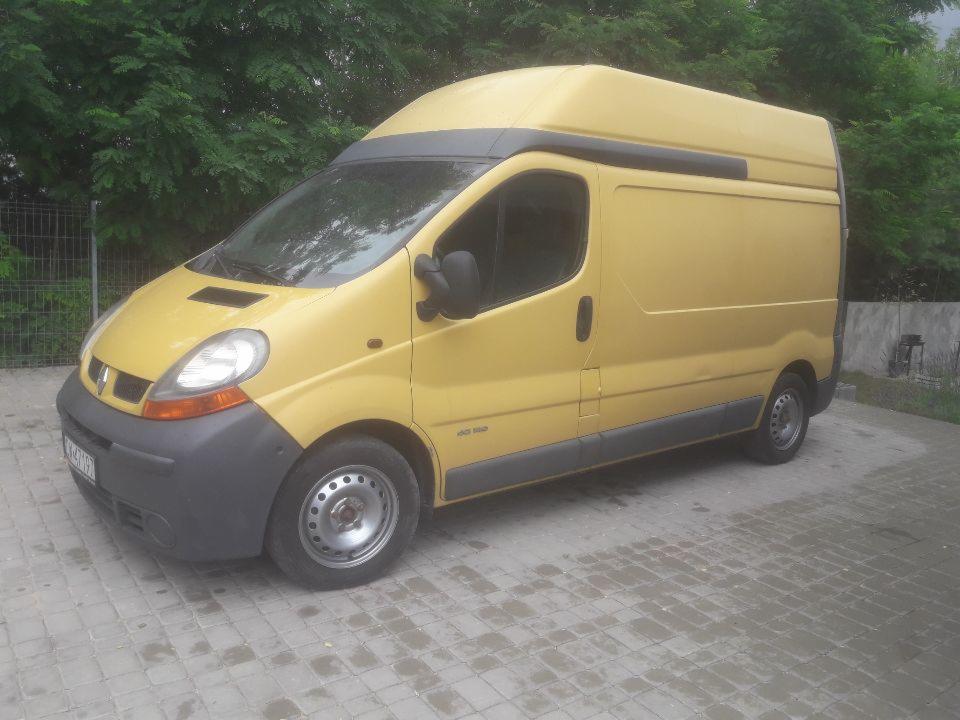 Żółty, wysoki bus Renault Trafic na kostce brukowej, z zielonym tłem drzew. Widoczny z boku, lekko od przodu.