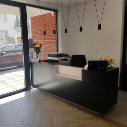 Max-Meble Design Robert Grabowski - Czarna lada recepcyjna z białym akcentem, oświetlona wiszącymi lampami, z widokiem na ulicę przez szklane drzwi i parkujący samochód.