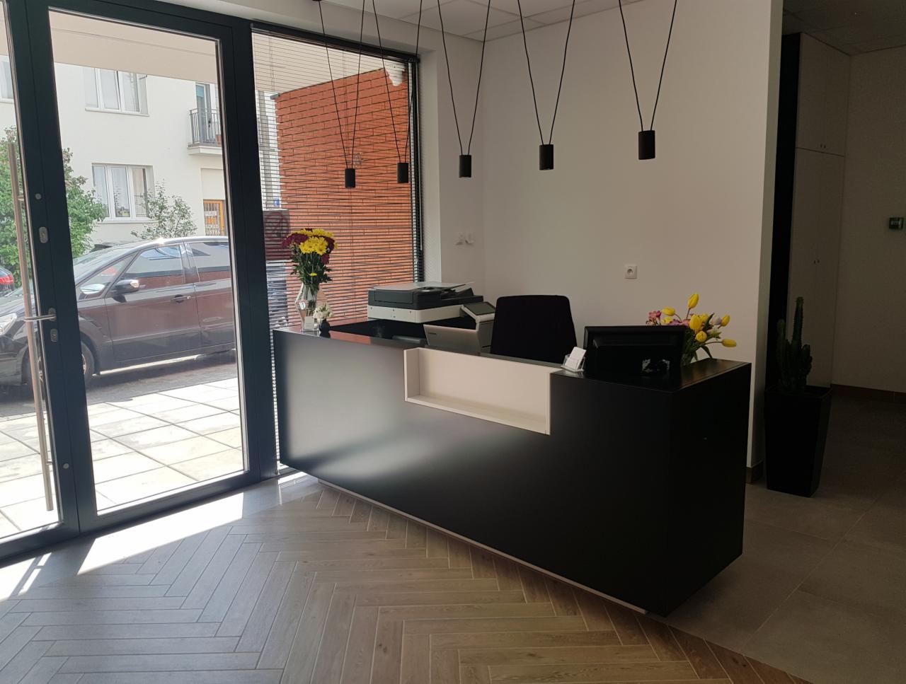 Czarna lada recepcyjna z białym akcentem, oświetlona wiszącymi lampami, z widokiem na ulicę przez szklane drzwi i parkujący samochód.