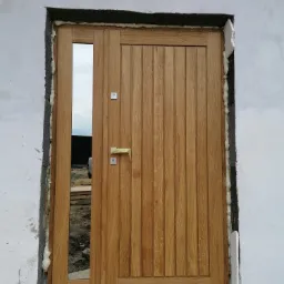 Montaż zewnętrznych drzwi drewnianych z pionowymi panelami i bocznym przeszkleniem, widoczne pianowanie ościeżnicy w surowym murze.