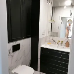 Nowoczesna łazienka z czarnymi szafkami, marmurowymi płytkami i złotymi akcentami, w lustrze odbija się osoba.