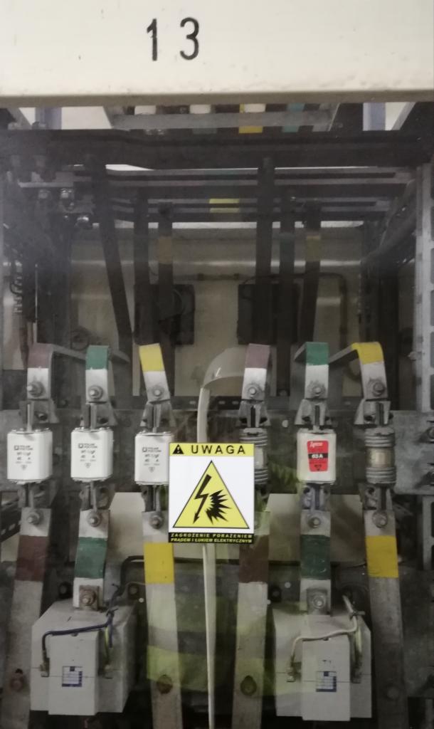 Rozdzielnica elektryczna z bezpiecznikami, oznaczeniami kolorystycznymi i tabliczką ostrzegawczą przed porażeniem prądem, numer identyfikacyjny '13' u góry.