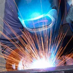 Intensywne iskry podczas łączenia metalowych elementów, ochronna maska spawalnicza z niebieskim odblaskiem, widoczne rękawice ochronne i perforowane tło.