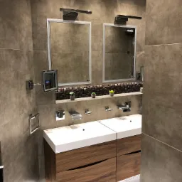 Nowoczesna łazienka z dwoma umywalkami na drewnianej szafce, podświetleniem LED przy podłodze, dwoma lustrami z oświetleniem i dekoracyjną półką z mozaiki i roślinami.