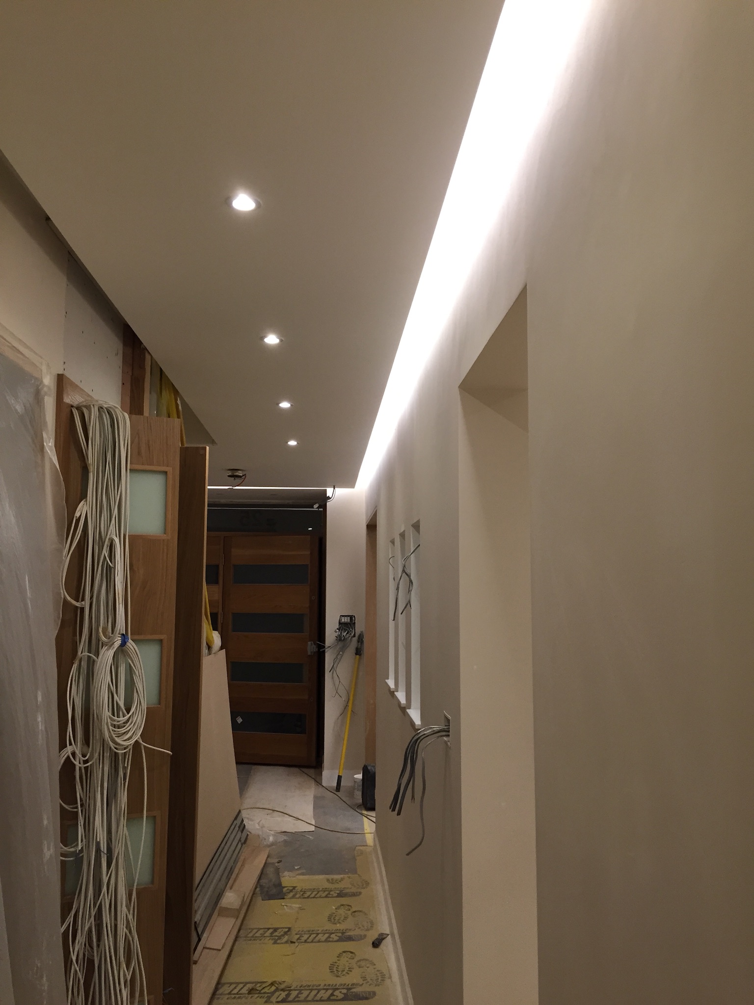 Korytarz w trakcie remontu z widocznymi kablami, punktowymi i liniowymi źródłami światła w suficie, oraz częściowo zamontowaną stolarką drzwiową.