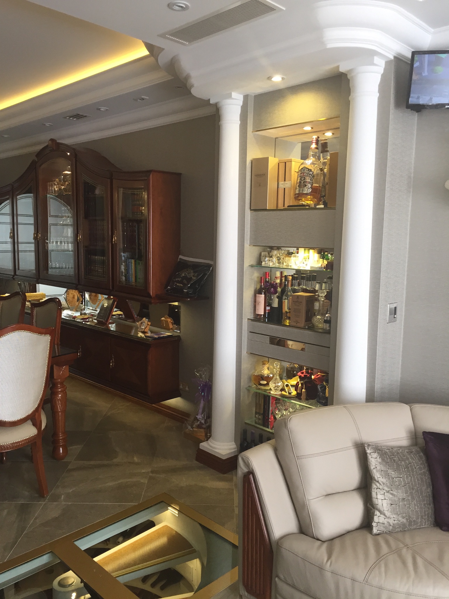 Luksusowe wnętrze salonu z podświetlaną wnęką, w której znajdują się szklane półki z kolekcją alkoholi i szkła, widoczne kolumny architektoniczne i fragment sofy.