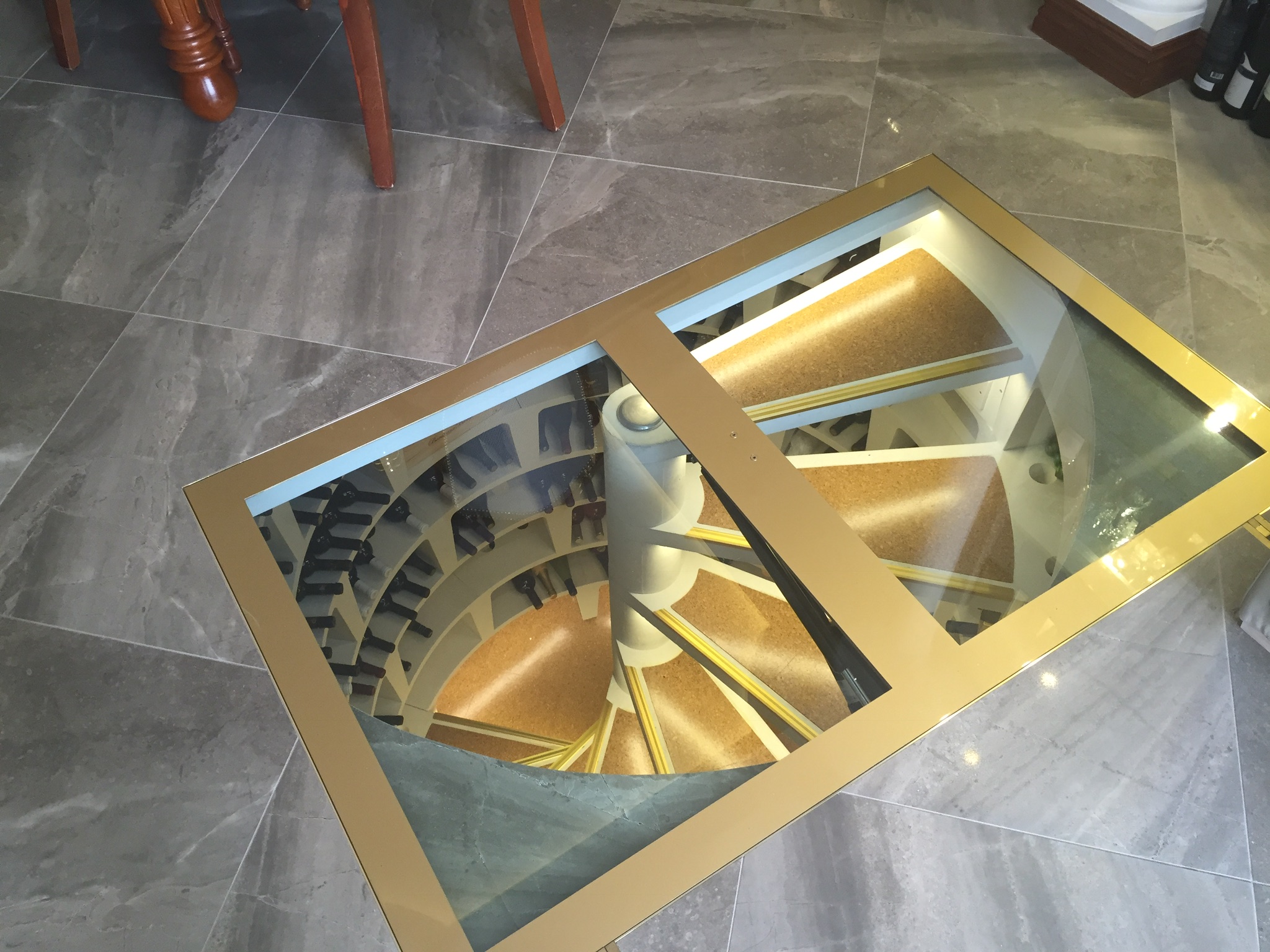 Szklana podłoga z widokiem na spiralną piwniczkę na wino, widoczna z góry, z fragmentem podłogi z płytek ceramicznych i nóg krzesła w tle.