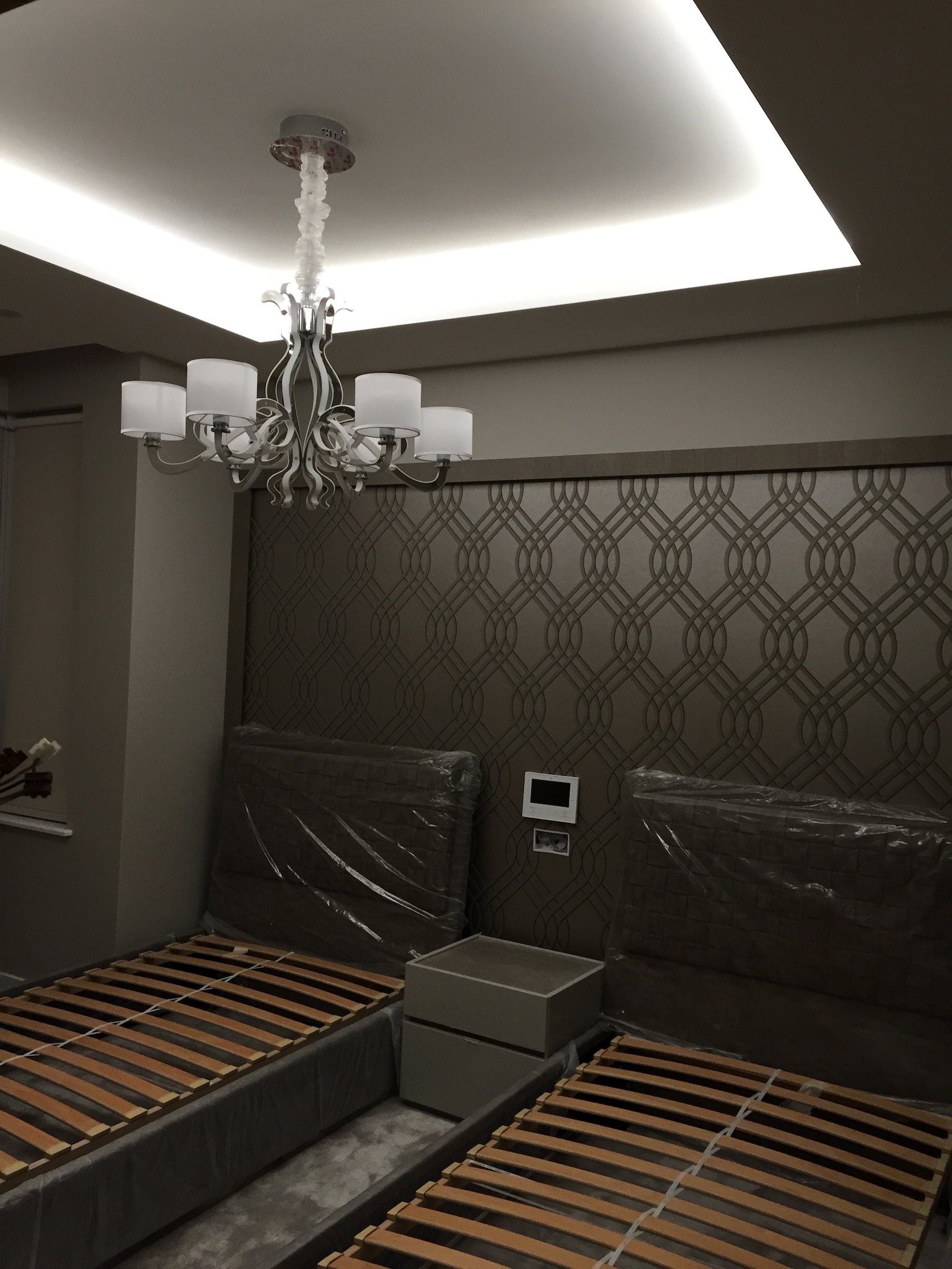 Nowoczesny pokój z dwoma łóżkami w folii ochronnej, nocną szafką i ozdobnym oświetleniem sufitowym LED oraz żyrandolem z abażurami, jeszcze w zabezpieczeniach.