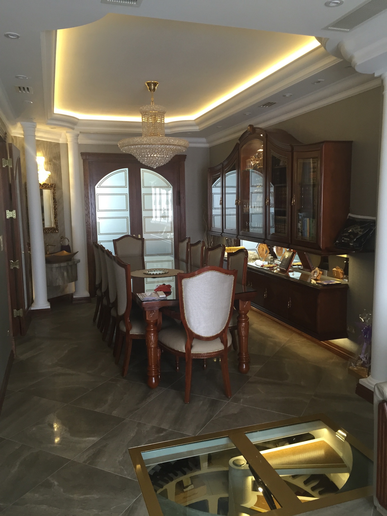 Elegancka jadalnia z kryształowym żyrandolem, podświetlanym sufitem i przeszkloną podłogą odsłaniającą piwniczkę z winami.