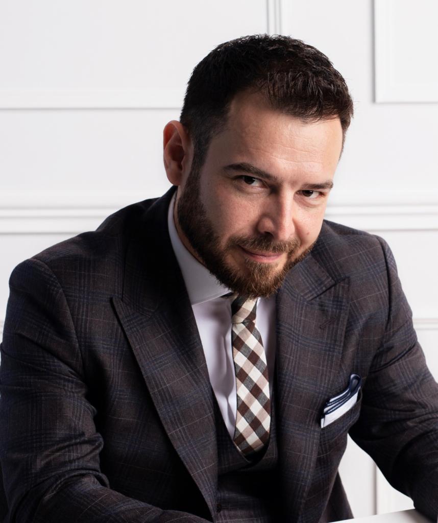 Portret brodatego mężczyzny w eleganckim, brązowym garniturze w kratę i krawacie w brązowo-białą kratę na tle białej ściany.