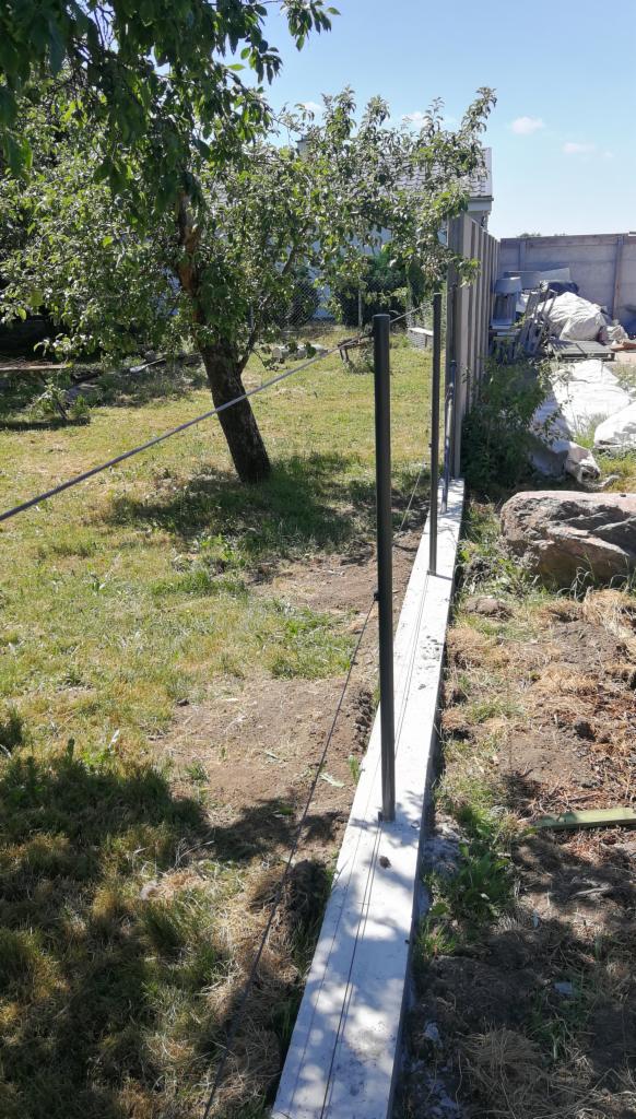 Budowa ogrodzenia panelowego: betonowa podmurówka z zamontowanymi słupkami na trawiastym terenie z drzewem w tle.