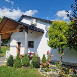 Biały dom z klimatyzatorem na ścianie, drewnianą wiatą wspartą na kolumnach, zadbanym ogrodem i niebieskim niebem w tle.