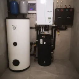 Kompletna instalacja grzewcza w kotłowni: biały zasobnik Kospel, czarny zbiornik, pompa ciepła Ecodan i moduły Buderus na tle szarych płytek.