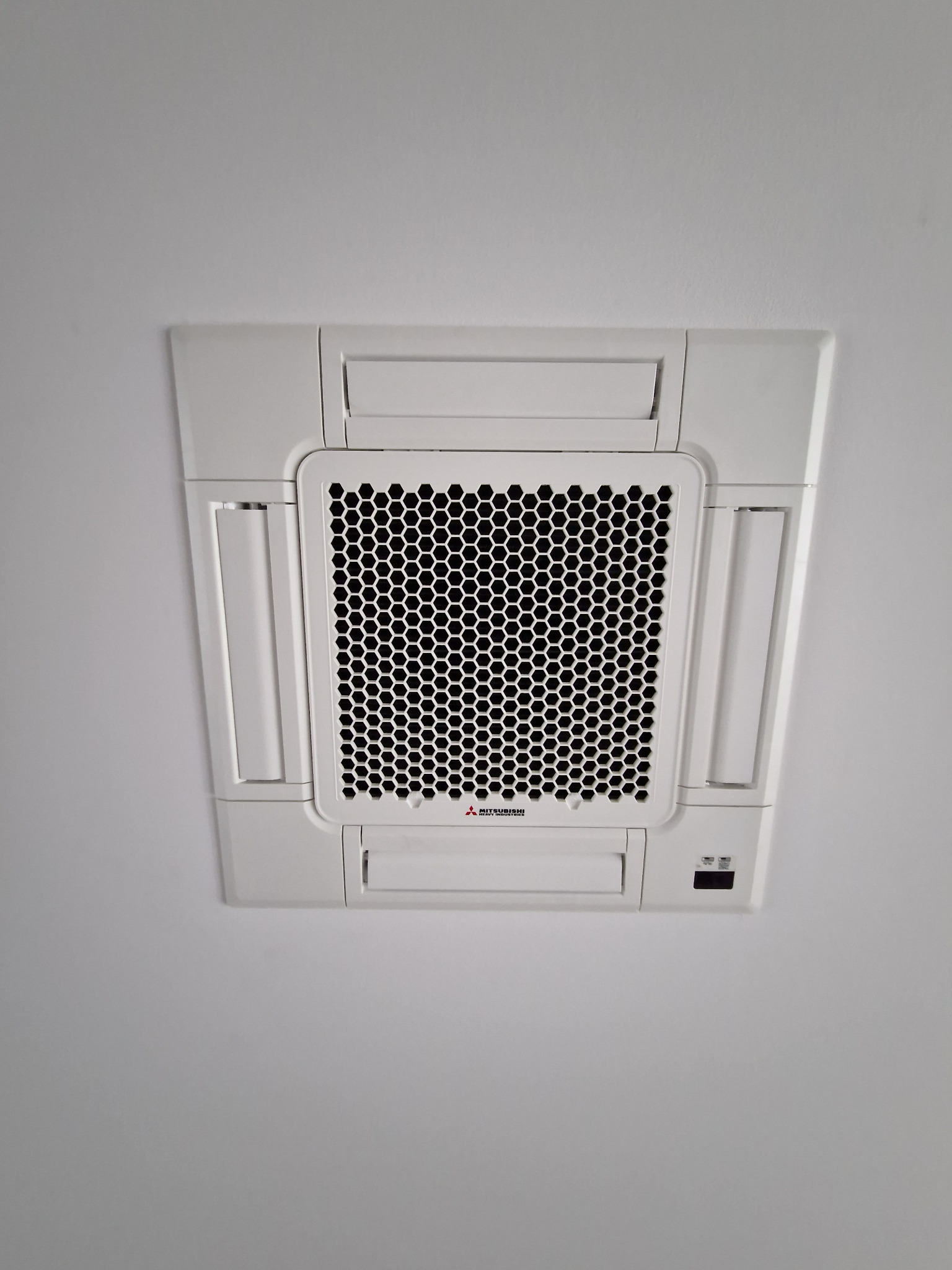 Biała, sufitowa klimatyzacja kasetonowa Mitsubishi Heavy Industries z kratką o strukturze plastra miodu, zamontowana na gładkim, białym suficie.