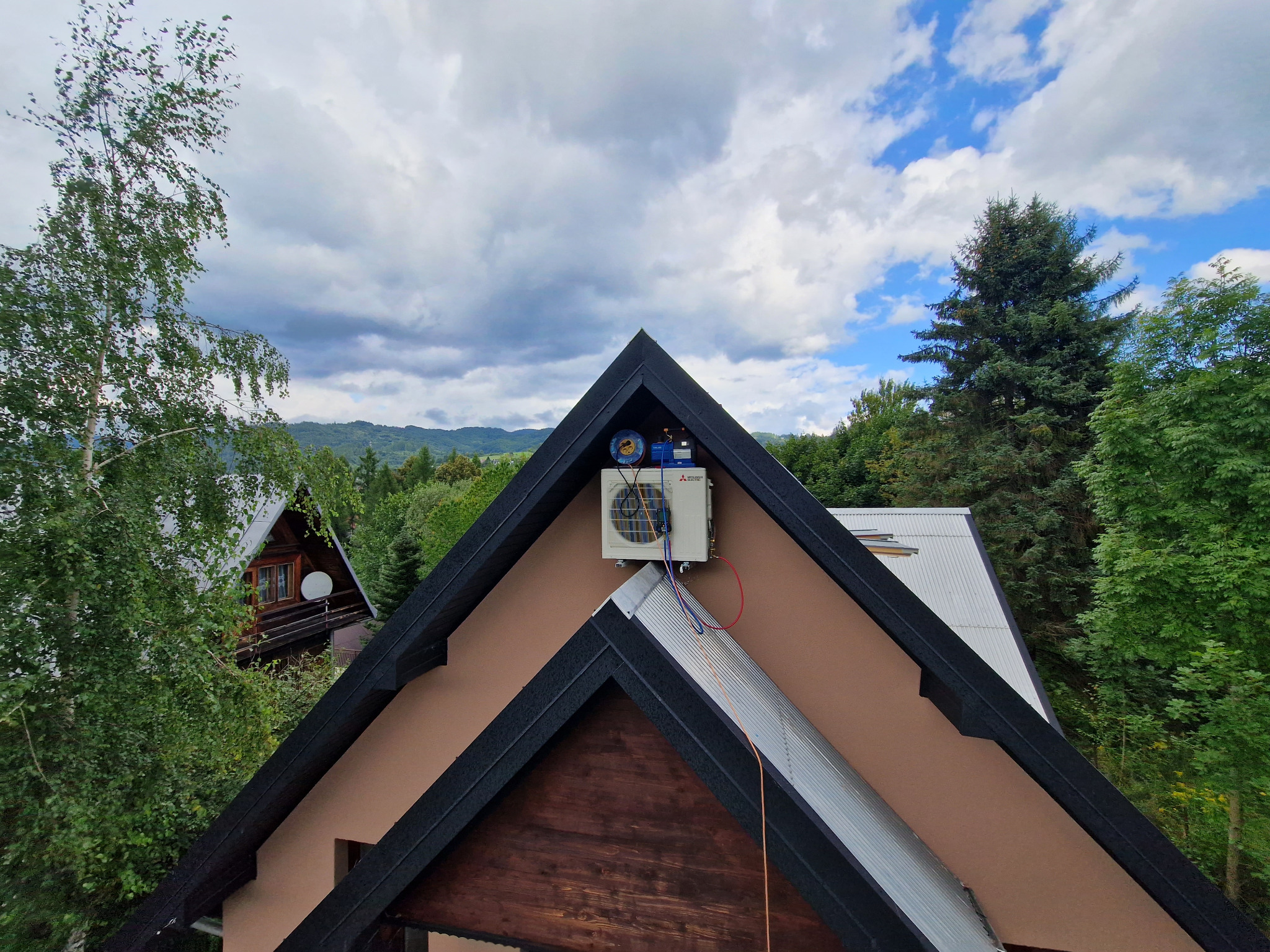 Zewnętrzna jednostka klimatyzacji Mitsubishi Electric zamontowana na ścianie domu z widocznymi przewodami, na tle lasu i pochmurnego nieba.