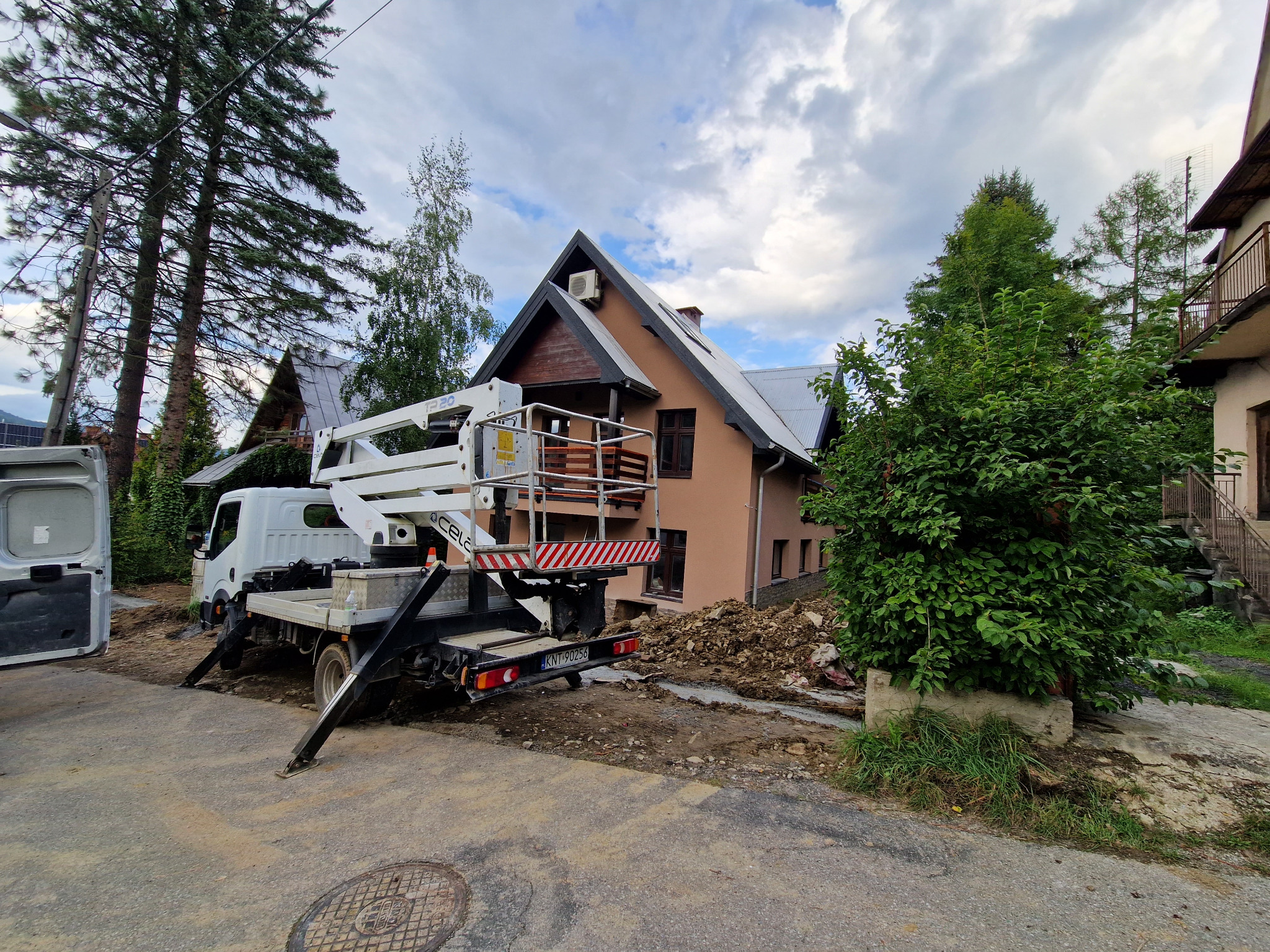 Montaż klimatyzacji na zewnątrz domu z użyciem podnośnika koszowego; widoczna jednostka zewnętrzna na ścianie budynku.