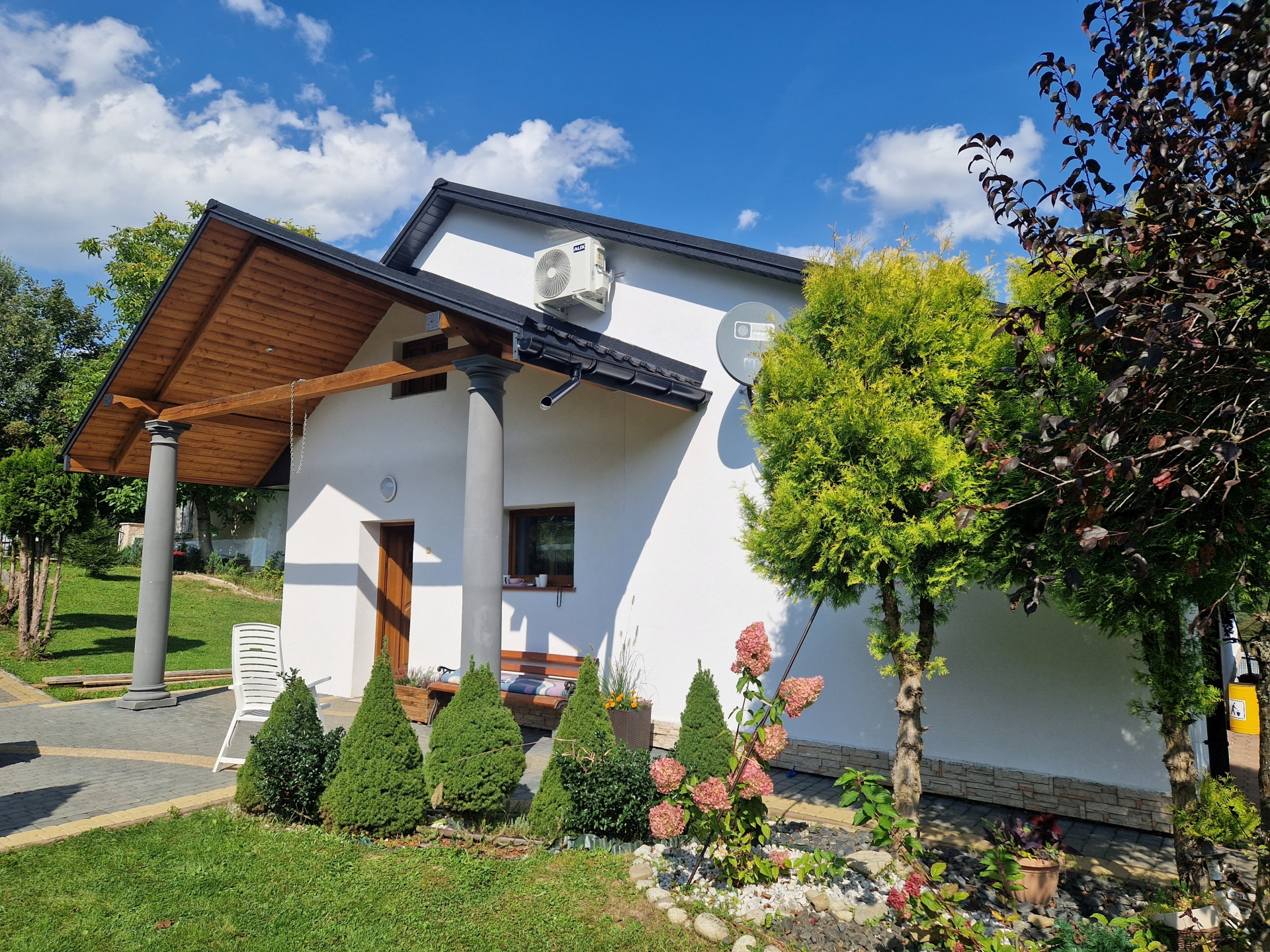 Biały dom z klimatyzatorem na ścianie, drewnianą wiatą wspartą na kolumnach, zadbanym ogrodem i niebieskim niebem w tle.