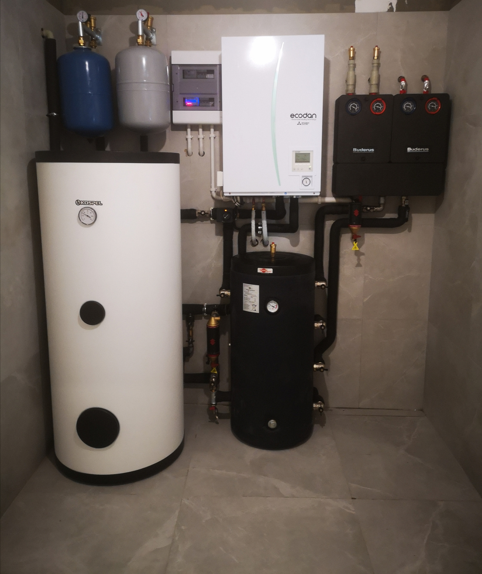 Kompletna instalacja grzewcza w kotłowni: biały zasobnik Kospel, czarny zbiornik, pompa ciepła Ecodan i moduły Buderus na tle szarych płytek.