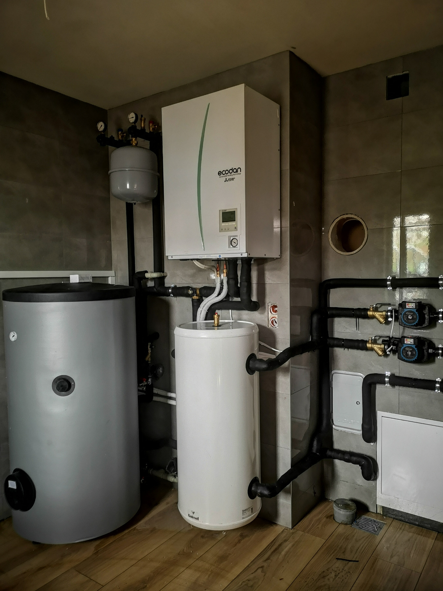 Zainstalowana pompa ciepła marki Ecodan w kotłowni w Szczawnicy, widoczne zbiorniki buforowe i rozdzielacze, rury w izolacji, szara płytka na ścianach i drewnopodobna podłoga.