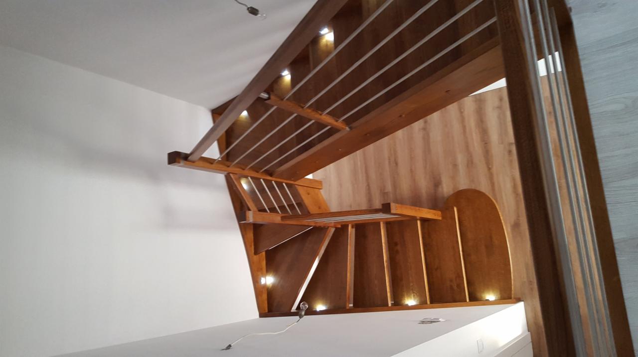 Nowoczesne schody z drewna z oświetleniem LED wbudowanym w konstrukcję, widoczne białe ściany i drewniana boazeria z zaokrąglonym wycięciem.