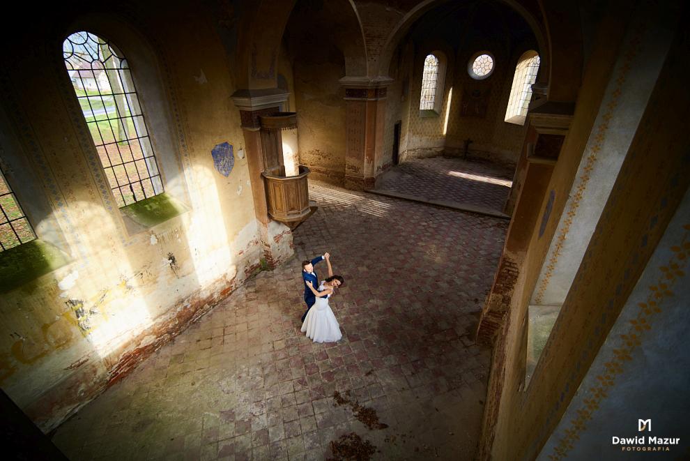 Para taneczna w ruinach kościoła, mężczyzna w granatowym garniturze prowadzi kobietę w białej sukni ślubnej, widok z góry na ceglaną posadzkę i promienie słoneczne wpadające przez okna.