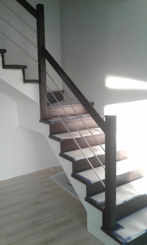 Nowoczesne schody z ciemnego drewna z metalowymi barierkami, zabezpieczone folią malarską podczas remontu, widoczne z dołu, z jasnym światłem padającym na ścianę.