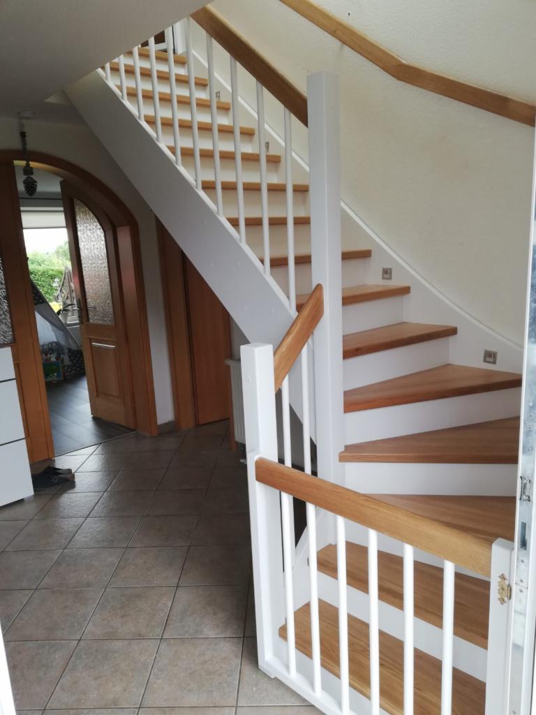 Jasne schody drewniane z białymi balustradami i drewnianymi poręczami, widoczne z dołu, z fragmentem otwartego wejścia i korytarza w tle.